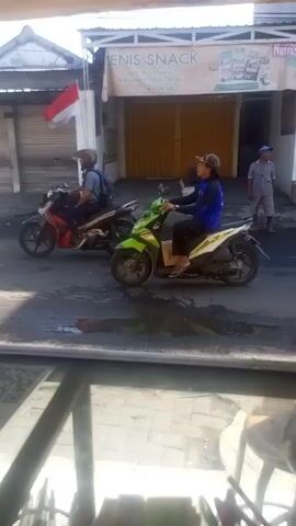 The video shows a street scene outside a shop called "DENIS SNACK." Two people carry a large black rectangular object into the shop. Several motorbikes pass by on the street. There is a small Indonesian flag waving in front of the shop.  A man in a blue shirt and a cap stands in front of the shop watching. The view is filmed from inside a building looking through a window.  The street is wet.