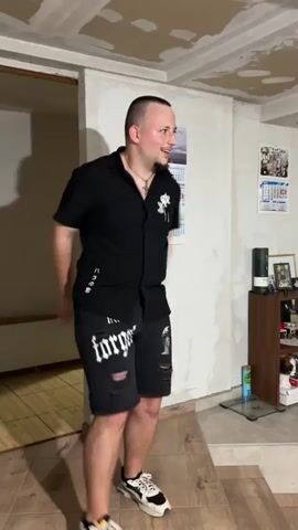 A man in a black shirt and ripped shorts dances enthusiastically in a room with unfinished ceilings. He then turns and faces two other men, one of whom is sitting on a ledge and wearing a black t-shirt and a baseball cap. The dancing man places his hands on the ledge, and the seated man also places his hands on the ledge. The third man, wearing a black shirt and shorts, joins them and places his hands on the ledge as well. The dancing man then pushes off the ledge. The scene takes place in what appears to be a living room with a wood-burning stove, shelves filled with items, and a mirror reflecting the room.