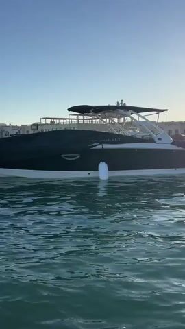 The video is a vertical, first-person perspective of a boat on the water. The boat is mostly covered by a dark tarp, with the brand "COBALT" visible. The stern of the boat shows a white swim platform and a white propeller. The water is clear and blue-green with small waves. In the background, a dock with floating barriers is visible, and behind that, a parking lot with several cars and some palm trees. The sun is setting, casting a warm glow on the scene. The sky is a clear, light blue.