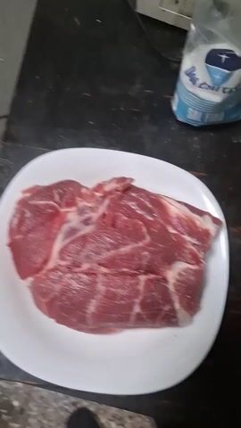 Here's a brief description of the video:

A raw cut of meat rests on a white, square plate on a black countertop. A hand holds a small, round container with a white lid and shakes a mixture of spices onto the meat. A bag of salt is visible in the background.
