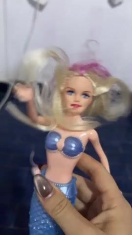 Here is a description of the video:

A hand is holding up a Barbie mermaid doll. The doll has blonde hair with a pink streak, blue eyes, and is wearing a blue seashell bra and a blue mermaid tail. The fingernails of the person holding the doll are long and painted with a shimmery brown color. The background is blurry, but there is a dark blue surface and a white object out of focus. The hand moves the doll around, showing its face from different angles.