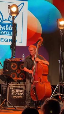 A man plays a double bass on a stage. He is wearing a dark short-sleeved shirt and jeans. He is standing in front of a large speaker cabinet and a monitor speaker. A backdrop with colorful circles and a logo that includes a rabbit is behind him. There are stage lights on either side of him. A music stand is to his left. The audience is visible in the foreground.