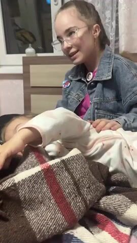 Here's a description of the video:

The video shows two young females sitting on a bed covered with a brown, red, and white checkered blanket.  A toddler with a ponytail is lying on the blanket, propped up on her elbows and looking to the left. She's wearing a white pajama top with a pink pattern. Behind the toddler is a teenager wearing glasses, a pink top, a denim jacket, and dark pants. The teen is sitting with her legs crossed and also looking to the left, seemingly in the same direction as the toddler.

The bedroom setting includes a light wood-colored headboard and a window with white frames. A baby bottle is visible on the windowsill. The lighting in the room appears to be soft, possibly from overhead lighting and natural light coming through the window.