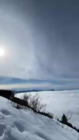 The video shows a winter landscape with a mountain ridge covered in snow. The view extends over a sea of clouds, with the tops of distant mountains visible in the background. The sky is a pale blue with thin, streaky clouds. In the foreground, there are snow-covered slopes with small trees and shrubs. The video slowly pans across the landscape.