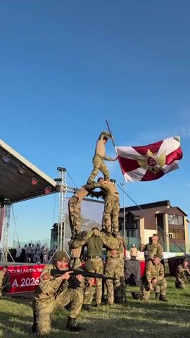 The video shows a group of people in military uniforms forming a human pyramid outdoors. One soldier stands atop the pyramid, holding a flag. Another soldier kneels in front, aiming a rifle. In the background, there's a stage, a building, and a group of spectators. The sky is clear and blue. There are signs in the foreground written in Russian and including the number 2026.