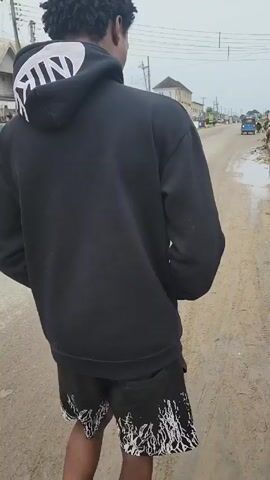 The video shows a man with dark, curly hair, wearing a black Nike hooded sweatshirt and black shorts with white designs, walking down a paved road. The camera follows him from behind.  He is joined by another man walking beside him. Buildings, parked cars, and power lines are visible in the background, giving the impression of an urban or suburban environment. The day appears overcast.