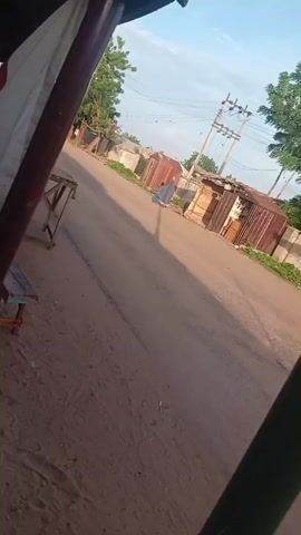 A person is walking on a dirt road. There is a bus stop with a bench and a sign. The sign has red and white text, but it is too blurry to read. There are buildings and trees in the background. The sky is blue with some clouds. The video is shaky and tilted.