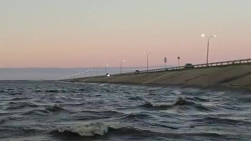 A wide shot captures a body of water with small waves rolling in. A concrete embankment lines the right side of the water, topped by a road with a railing. Streetlights are spaced along the road, and a few vehicles are visible driving on it. The sky above is a soft gradient of pink and pale blue, suggesting either dawn or dusk. The overall scene is tranquil and expansive, with the natural element of the water contrasting with the man-made structures.