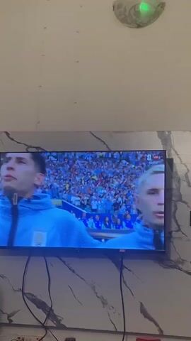 The video shows a television screen mounted on a wall. The television is displaying a soccer match. The screen shows several male soccer players, some of whom are wearing light blue jerseys. One player in the foreground is looking up and to the left. The background shows a stadium filled with spectators. The wall the television is mounted on has a marble-like pattern. There is a light fixture on the wall above the television, and cables are visible below the television. The video is taken from a slightly angled perspective.