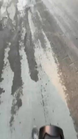 Here is a concise description of the video:

The video shows a dilapidated road with many potholes and tire tracks. Several vehicles, including vans, trucks, and cars, are parked on the side of the road. A group of people is standing near the vehicles, seemingly observing something in the distance. Smoke is visible rising into the air behind them, suggesting a fire or some other form of combustion. The sky is clear, with the sun rising in the distance.
