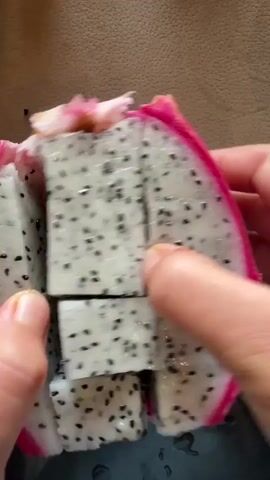 The video shows a close-up of a person's hands slicing a dragon fruit. The fruit has white flesh with black seeds and a pink rind. The hands are shown cutting the fruit into several pieces. The background is out of focus and appears to be a brown surface. The lighting is bright.