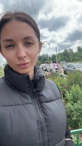 The video begins with a close-up shot of a young woman from the chest up, facing the camera. She has dark hair pulled back, light-colored eyes, and a neutral expression. She is wearing a black puffer vest zipped up over a black shirt. The background is slightly out of focus but shows a paved area, some greenery, and parked cars, suggesting an outdoor urban or suburban setting. The camera movement is subtle, with slight shifts in angle and focus, giving the impression of a selfie video. The lighting is natural, indicating it's daytime. The woman's head slightly tilts from side to side throughout the short clip, and she blinks a few times. The overall impression is a casual, brief recording.