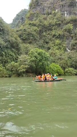 A group of people is riding in a long, narrow boat on a river. The boat is green and has the number "307" painted on its side.  Most of the passengers are wearing orange life jackets and using oars to row. The boat is moving through the water. Two women wearing traditional conical Asian hats are also rowing. The river is green and surrounded by lush green hills and trees.  The scene suggests a tourist activity or a guided tour through a natural waterway.