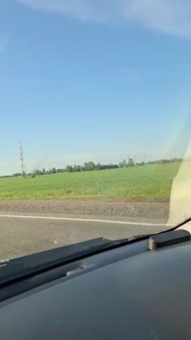A view from inside a vehicle looking out the windshield. Outside, a bright blue sky with a few wispy clouds stretches over a vast, green field. Power lines are visible stretching across the horizon, supported by tall, lattice-style towers. The field appears to be a crop of some kind, with some trees dotting the landscape in the distance. The foreground shows the car's dashboard and windshield wiper. The overall impression is a scenic drive through the countryside on a clear day.
