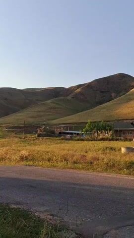 A serene landscape unfolds with rolling hills under a clear blue sky. In the middle ground, a rustic house with a brown roof sits amidst dry, golden grass. A paved road runs in the foreground, leading the eye towards the tranquil scene.