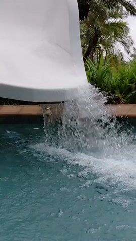 A white water slide curves down towards a swimming pool. Water cascades from the end of the slide into the pool, creating ripples and splashes. In the background, lush green foliage, including palm fronds, is visible. The lighting suggests an overcast or indoor setting. A person's legs are briefly visible as they appear to be moving under the slide.