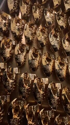 The video shows a display of identical golden crab-shaped objects, possibly trophies or awards, arranged in a grid pattern. Each item features a detailed crab claw design, seemingly made of shiny metal. A band, adorned with dollar sign symbols, is attached to each crab claw. The overall scene has a luxurious and opulent feel, due to the use of gold color and the repetitive arrangement. The lighting appears to be indoor, reflecting off the metallic surface of the objects. There are no people visible in the video, and the focus remains solely on the display of golden crab claws.