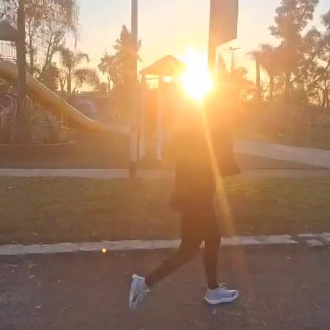 A person is running on a paved path at dusk, with a playground visible in the background. The sun is setting, casting a warm glow on the scene. The person is wearing athletic clothing and appears to be moving at a moderate pace. The path is surrounded by grass and trees, and there is a slight incline. The overall mood is peaceful and serene, with a sense of movement and activity.