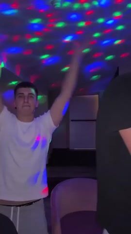 The video shows a young man dancing in a room lit with colorful disco lights. He's wearing a white t-shirt and gray sweatpants, moving his arms and body to the music. The camera pans around the room, showing other young people sitting at tables, some watching and some on their phones. There are bottles of Coca-Cola and cans on the tables. The room has a dark, club-like atmosphere with patterned wallpaper.