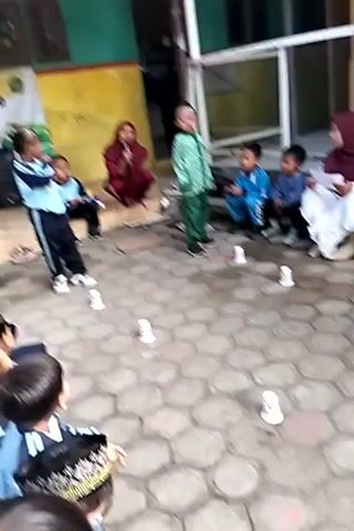 A group of young children and adults are gathered on a paved outdoor area with a honeycomb pattern. The children appear to be participating in a game or activity involving small white cups placed on the ground. Some children are standing, some are sitting, and others are on their hands and knees. Two women in headscarves are observing the activity, one appears to be speaking into a microphone or holding a paper. One child, dressed in a green patterned outfit, is standing in the center. Another child in a blue tracksuit is on the left, facing the group. The adults and some of the children are seated on a raised platform in the background. The scene appears to be an outdoor event, possibly a school or community gathering.