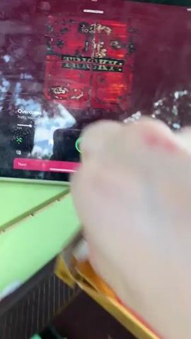 The video shows a close-up of a table with a tablet playing music, a pack of cigarettes, a lighter, and a book. A person's hand is seen reaching for the cigarette pack. The tablet displays album art and playback controls. The lighter is ornate, with a dragon design and Chinese characters. The cigarette pack is red and gold, also with Chinese text. The background is blurry, suggesting an outdoor setting with trees and foliage. The camera then pans out slightly to reveal more of the table and the person's lap, which is covered by a floral-patterned fabric.