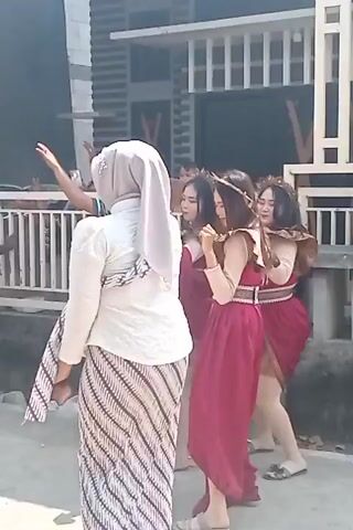 This is a video of an outdoor event. In the foreground, a person wearing a white hijab and a patterned skirt is walking away from the camera. Behind them, three people are standing, all wearing similar maroon dresses and some sort of headwear. One person in the background, wearing a blue shirt and a cap, appears to be holding a phone and taking a picture. The environment seems to be a street or pathway with a building in the background, possibly a house with a decorative white fence. The lighting suggests it is daytime.