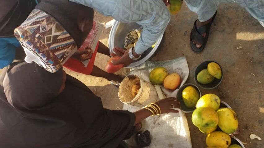 The scene presents a high-angle view of a group of people engaged in a transaction involving mangoes.  A woman wearing a black hijab and multiple gold bangles is seated on the ground, examining a mango in her hands.  She's situated near a collection of yellow and green mangoes arranged in several small bowls and on a cloth.  A man with a patterned cap stands near her, while another man in a light blue shirt reaches into a large silver basin full of mangoes, offering a red mango to the woman. A child wearing red shorts is also present in the scene.  The ground is mostly a mixture of dirt and concrete, littered with small scraps of organic material. A woven basket sits near the mangoes, and a pair of sandals can be seen near the basin. The scene appears to be outdoors under natural lighting conditions.