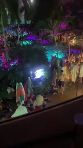 The video shows a crowded indoor water park at night. People are gathered in a large pool, with palm trees and other tropical plants interspersed throughout the area. The pool is illuminated with colorful lights, predominantly pink and green. A glass ceiling covers the entire space. In the foreground, there are towels and personal belongings scattered on the ground near a walkway, with several people standing around. The music is loud and upbeat. The camera slowly pans upwards, revealing more of the pool and the large crowd.