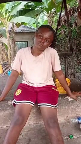The video shows a person sitting on a concrete step in an outdoor area. The person is wearing a light pink t-shirt and maroon shorts with white trim, and sandals. The person has dark skin and braided hair. They are leaning to the side with their head tilted, and their eyes are closed. Behind them, there are banana trees and a small building. There are also several yellow and blue containers scattered around. To the right, there is a small metal bowl, a water bottle, and another container on the concrete step. In the background, there appears to be someone working with metal. The camera moves slightly during the video.