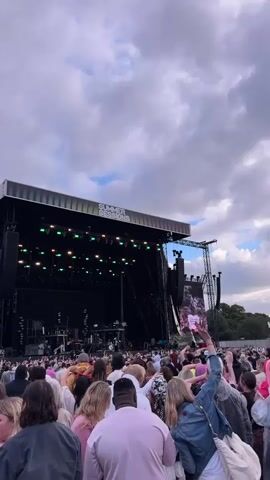 The video shows a large outdoor music festival crowd gathered in front of a stage. The stage is large and lit with green lights. A banner at the top of the stage reads "SUMMER SESSIONS". Several large speakers are visible on either side of the stage. The crowd is diverse, with people of all ages and ethnicities, many of whom have their arms raised in the air. The sky is cloudy with patches of blue. The camera is held at eye level, suggesting the viewpoint of someone in the crowd.