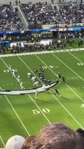 The video shows a football game in progress from a high vantage point, likely within the stadium. The field is green with yard lines and markings. Players in both white and black uniforms are positioned on the field, seemingly mid-play.  Spectators fill the stadium seats, with many wearing team colors. The day appears sunny. The focus is on the players and the ongoing play on the field.