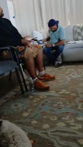 The video shows a group of people sitting in a living room. One man is sitting in a gray chair, wearing a black hoodie, patterned shorts, and orange sneakers. Another man is sitting on a white couch, wearing a blue baseball cap, light blue shirt, and jeans, looking at a phone. A third man is sitting on the same couch, wearing a black shirt and jeans. A woman is sitting on the couch wearing a black and white tank top and leggings. A dog is laying on the rug. There is a coffee table with a keyboard and mouse on it. The room has white curtains and a patterned rug. A TV is visible in the background. All the people are looking at their phones except for the man in the black hoodie, who is speaking.