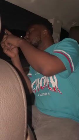 An African-American man with a short beard is sitting in the back of a car. He is wearing a light blue t-shirt with white and red accents and light gray jeans. He is looking down at a phone in his hands. The car has dark leather seats and a sunroof. The video appears to be taken at night.
