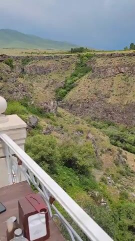 The video pans across a vast canyon. The canyon walls are steep and rocky, with vegetation growing in patches. The top of the canyon is flat and appears to be grassland. The sky is overcast with some clouds. There is a white railing in the foreground of the video.