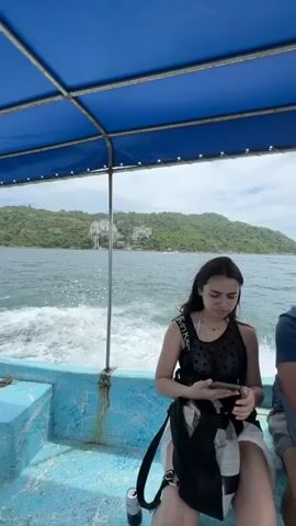 Four people are on a light blue boat with a dark blue canopy, moving across calm water. A young woman with dark hair, wearing a black mesh top over a white shirt and light shorts, is sitting and looking at her phone. To her right, a man with dark hair, wearing a blue t-shirt and light shorts, is looking forward with a light blue bag on his lap. Further back, another woman with dark hair, wearing a pink top, holds up a phone to take a picture of the coastal scenery. Another person, seen from behind wearing a white shirt and a brown-and-white patterned scarf, holds onto a metal support pole. The background features green, hilly coastline dotted with white buildings under an overcast sky. The boat generates a white wake behind it.