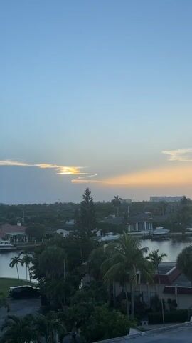 The video shows a wide shot of a suburban area with a body of water. The sky is blue with some clouds, and the sun is setting. There are houses and trees visible, as well as some palm trees. The sun's rays are visible through the clouds, creating a dramatic effect. The overall mood is peaceful and serene.