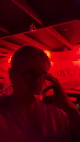 A person is shown in the center of the frame, illuminated in a strong red light, drinking from a glass. The person is wearing glasses and has short, light-colored hair. The background is dimly lit, suggesting a bar or club setting, with indistinct shapes of other people and furniture. The video is shot from a low angle, looking up towards the ceiling, which has a structured, grid-like design. There is a disco ball and some decorative elements hanging from the ceiling. The overall atmosphere is dark and vibrant, owing to the strong red lighting.