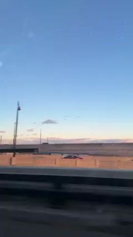 The video shows a view from inside a vehicle moving on a highway. The sky is blue with some light clouds, suggesting early morning or late afternoon. A bridge is visible in the background, with streetlights lining its sides. The vehicle is passing under metal structures, possibly for signage or support. In the distance, a construction crane is visible. Other vehicles can be seen on the adjacent road. The perspective is from the passenger side of the car, looking towards the right.