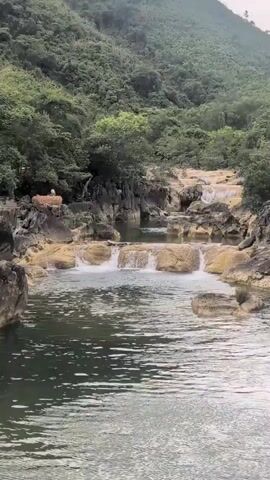 The video shows a natural scene with a river flowing through a rocky landscape. The river has several small waterfalls cascading over rocks. Lush green vegetation covers the surrounding hills. There are a few rocks visible in the foreground and midground, with some people standing on a rock formation to the left of the frame. The water is calm and reflects the surrounding greenery. The overall impression is one of a peaceful and scenic natural environment.
