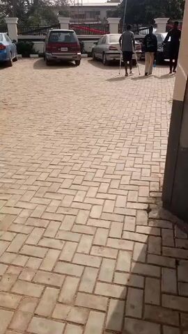 The video shows a paved parking lot with several cars parked in it. A man with crutches walks across the parking lot, accompanied by two other people. The parking lot is paved with light brown bricks in a herringbone pattern. A portion of a building wall is visible on the right side of the frame. The sky appears bright, suggesting it is daytime. The video is recorded from a slightly elevated position.