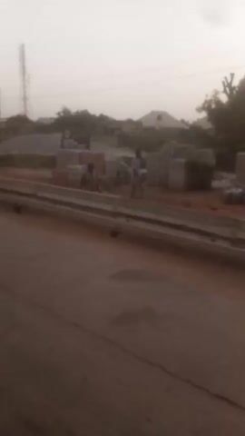 The video shows a view from a moving vehicle on a road. First, the view is of a construction site with piles of gravel and some building materials near the road. Next, a yellow motorized tricycle passes by. Then, two people in white robes are seen walking near the road. Finally, the view shifts to the perspective of a motorcycle rider heading down a road with other motorcycles and vehicles in the distance. The sky is overcast throughout the video.