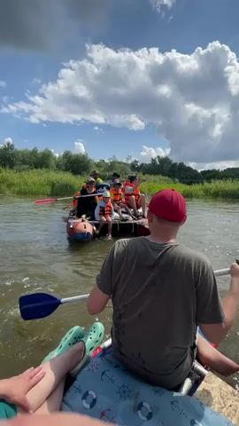 A group of people are on a raft on a river. The raft is made of inflatable tubes and has several people sitting and paddling. The people are wearing life jackets. The river is surrounded by trees and vegetation. The sky is blue with some clouds. The camera is positioned behind some of the people, looking towards the raft. One person in the foreground is wearing a red hat. The raft appears to be moving through the water.
