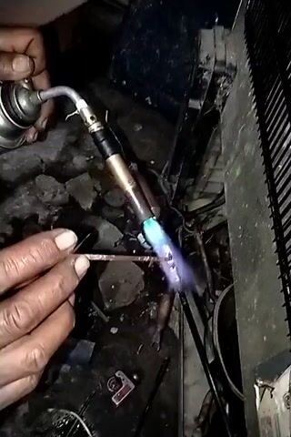 The video shows a close-up of a person's hands working on what appears to be a refrigeration unit. The person is holding a copper pipe in their left hand and a torch in their right hand. The torch is emitting a blue and pink flame, which is being used to heat the copper pipe. The pipe is connected to other components of the refrigeration unit, which are partially visible. The background is dark and cluttered, with debris on the floor. There is a black grid-like structure on the right side of the frame, which could be a condenser fan or grille. The overall impression is that of a repair or maintenance task being performed on a piece of equipment.