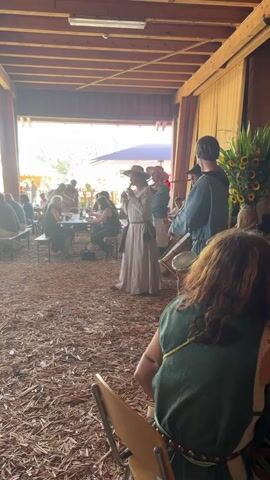 The video shows a performance in a barn-like structure with a wooden ceiling and walls. The floor is covered in wood chips. In the foreground, a person in a green tunic sits in a chair, facing away from the camera. In the middle ground, a group of people are performing music. One person in a white gown and hat is singing or speaking into a microphone. To their right, two people are playing instruments: one playing a flute-like instrument and another playing a drum. Several audience members are seated at tables in the background, some of whom are eating or drinking. Sunlight streams in from an open area in the background, illuminating the scene and creating a slightly hazy atmosphere.