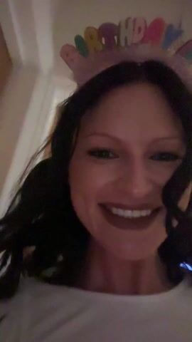 A woman with dark hair, a pink birthday crown, and a white shirt smiles at the camera. She is indoors. The camera moves slightly.
