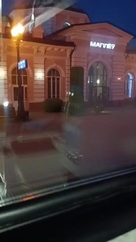 The video shows a reflection of a building with the word "МАГПЛЁ?" lit up in the center above the entrance. The building is light-colored with arched windows and is illuminated by lights. There are also illuminated street lamps visible. The reflection shows the building and the sky, which is dark blue, indicating it is nighttime. There are also blurred shapes visible in the foreground, suggesting the video was taken from inside a vehicle, possibly looking through a window. Some dark, vertical figures, possibly bushes or trees, are visible in front of the building, and there is a revolving door at the entrance.