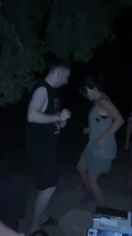 The video depicts a group of four people, two women and two men, standing outdoors at night, surrounded by trees. The lighting is dim, suggesting it might be from a campfire or other low light sources. The person on the far left, a man, is wearing a black tank top and black shorts. He appears to be talking to the woman next to him, who is wearing a light-colored top and light-colored shorts. She has dark hair and is smiling. Behind them, another man in a red floral shirt and shorts is visible. On the far right, another woman with long dark hair is wearing a blue and white patterned dress. She is talking to the woman in the light-colored top and shorts, and gestures with her right hand. The overall mood appears casual and friendly.