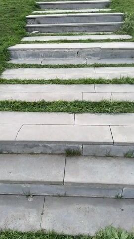 The video shows an upward-facing view of a set of concrete steps leading up a grassy hill. A single young girl is walking up the steps towards the top of the hill. At the summit, a large concrete statue of Jesus Christ stands prominently, surrounded by a number of smaller white crosses. The sky overhead is overcast and grey. The steps are interspersed with patches of grass growing between the concrete slabs.