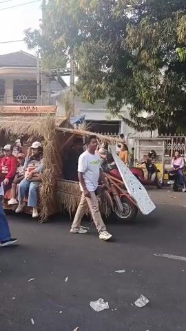 A short video shows a parade float constructed to resemble a traditional dwelling, possibly a hut. The float appears to be mounted on a vehicle, possibly a modified motorcycle with a trailer. It is decorated with dried straw or grass, giving it a rustic appearance. A sign is visible at the top of the float. Several people are seated on the float, including women and children. One person in the foreground, wearing a headscarf and loose pants, is walking beside the float. The parade is taking place on a paved street lined with buildings and trees. Other people can be seen observing the parade from the side of the street. The overall atmosphere seems festive.