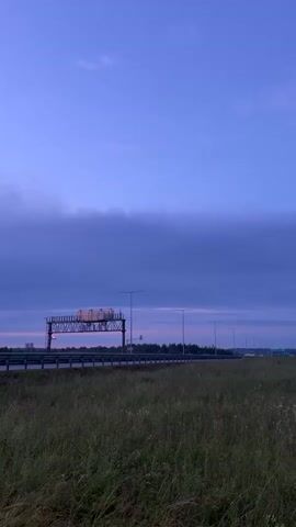 A static shot captures a rural landscape at dusk. A vast expanse of clear, pale blue sky dominates the upper half of the frame, gradually transitioning to a deeper, more muted blue towards the horizon. Wispy, dark blue clouds stretch across the sky, adding texture. In the middle ground, a highway stretches horizontally, bordered by a metal guardrail. To the left, a large, metallic gantry stands over the highway, supporting what appears to be an illuminated sign or message board, emitting a soft, white light, occasionally flashing with blue. Several streetlights, their tops obscured by the frame, are visible along the highway's edge. The foreground is filled with tall, green grass, dotted with small white wildflowers, swaying gently. The overall impression is one of quietude and the fading light of day.